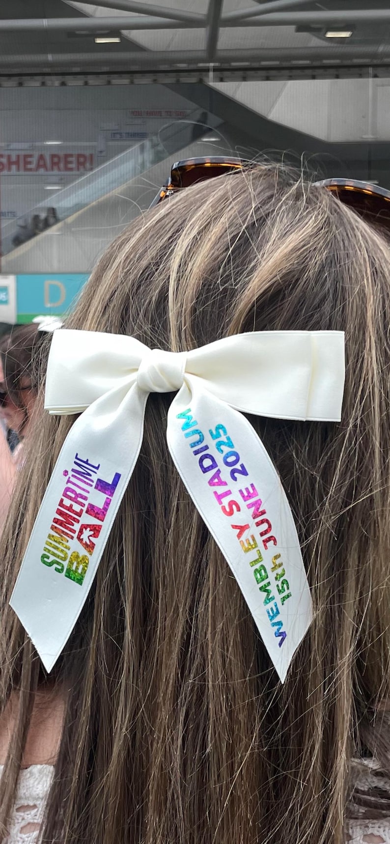 May include: A white hair bow with two long tails featuring colorful text. The left tail reads "SUMMERTIME BALL" in a multi-colored gradient, and the right tail reads "WEMBLEY STADIUM 15th JUNE 2025" in blue and green gradient lettering.