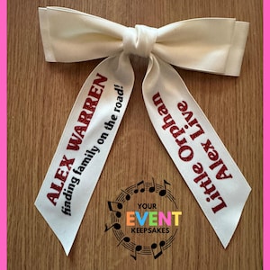 May include: A cream-colored fabric hair bow with two long tails. The left tail features red glitter text reading ALEX WARREN finding family on the road! The right tail features red glitter text reading Little Orphan Alex Live.