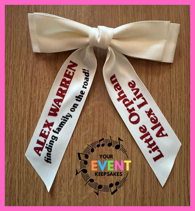 May include: A cream-colored fabric hair bow with two long tails. The left tail features red glitter text reading ALEX WARREN finding family on the road! The right tail features red glitter text reading Little Orphan Alex Live.