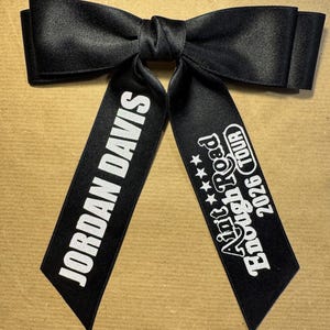 May include: A black satin hair bow featuring two long tails. The left tail displays the white text JORDAN DAVIS, while the right tail features the white text Aint Enough Road 2026 TOUR with four small stars above the tour name in a bold font.