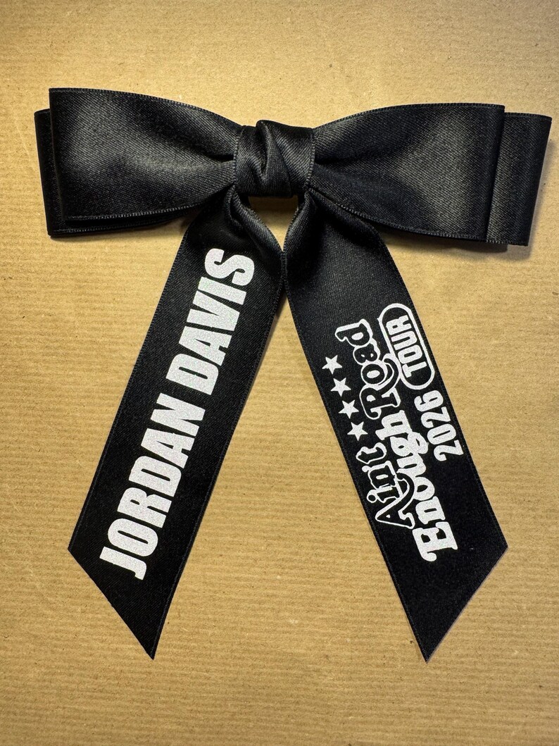 May include: A black satin hair bow featuring two long tails. The left tail displays the white text JORDAN DAVIS, while the right tail features the white text Aint Enough Road 2026 TOUR with four small stars above the tour name in a bold font.