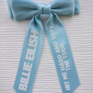 May include: A light blue satin hair bow with two long tails. The left tail features the white text BILLIE EILISH, and the right tail features the white text HIT ME HARD AND SOFT THE TOUR. The bow has a smooth, shiny finish and a central knot.