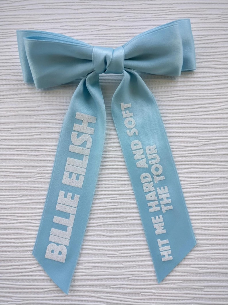 May include: A light blue satin hair bow with two long tails. The left tail features the white text BILLIE EILISH, and the right tail features the white text HIT ME HARD AND SOFT THE TOUR. The bow has a smooth, shiny finish and a central knot.