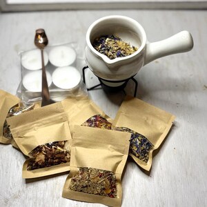 May include: A white ceramic oil warmer with a handle, filled with dried herbs and flowers, sits on a black metal stand. Several small paper pouches filled with herbs and flowers are scattered around. A small spoon and tea lights are also present.