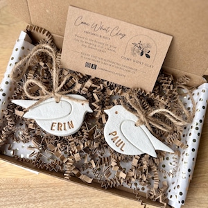May include: A gift box contains two white clay bird ornaments with the names "Erin" and "Paul" embossed in gold. The birds are tied with twine and rest on brown paper filler. A card with the text "Come What Clay" is also in the box.