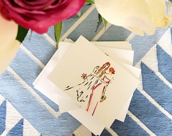 Puede incluir: Una pila de tarjetas blancas con una ilustración dibujada a mano de dos personas con atuendos de boda abrazándose. La ilustración está en tonos de rosa, naranja y amarillo. Las tarjetas están sobre una superficie con estampado azul y blanco con dos rosas, una rosa y una blanca.