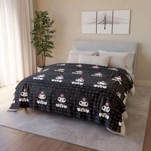 May include: A black blanket with a repeating pattern of a cartoon character with a pink skull and white details. The blanket is on a bed with white pillows and a grey headboard. The room has a neutral colour scheme.