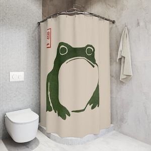 May include: Beige shower curtain featuring a large green frog illustration. The frog has a minimalist design with large eyes and a simple outline. A red square with Japanese text is in the upper left corner.