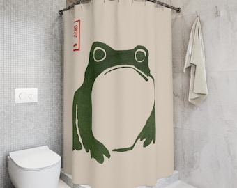 May include: Beige shower curtain featuring a large green frog illustration. The frog has a minimalist design with large eyes and a simple outline. A red square with Japanese text is in the upper left corner.
