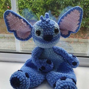 Peut inclure: Une peluche bleue faite à la main au crochet avec de grandes oreilles, des yeux noirs et des accents bleu foncé. La peluche est assise sur une surface blanche devant une fenêtre. Les oreilles sont rose clair à l'intérieur.