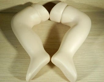 May include: Two pale, porcelain doll legs are positioned together, forming a heart shape. The legs are smooth and have a slight curve, with detailed feet. The background is a light wood grain.