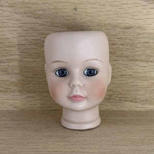 May include: A vintage doll head with a porcelain finish. The doll head has blue eyes, delicate eyelashes, and rosy cheeks. The lips are painted a soft pink. The head is a light beige color and has a smooth texture.