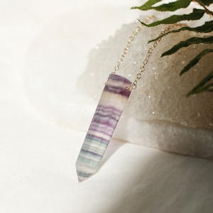 Puede incluir: Un collar de cadena dorada con un colgante de cristal de fluorita morado, verde y blanco. El colgante tiene forma de punta y una textura natural estriada.