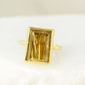 Pode incluir: Anel de ouro com uma pedra de quartzo transparente retangular com inclusões douradas. A pedra está engastada em uma moldura de ouro.