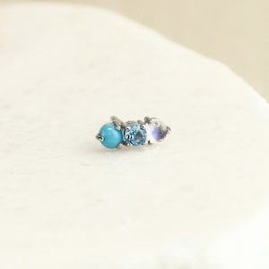 May include: A silver nose stud with three gemstones: a turquoise, a blue topaz, and a moonstone.