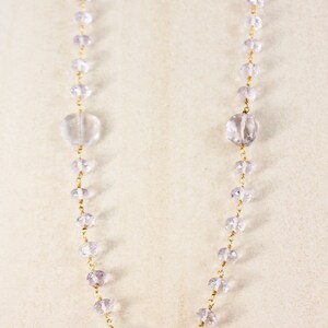 May include: A long, delicate necklace made of clear and pale purple faceted beads strung on a gold chain. The necklace features larger, clear faceted beads at the ends and in the center.
