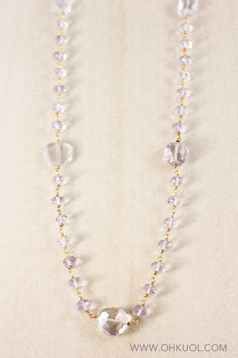 May include: A long, delicate necklace made of clear and pale purple faceted beads strung on a gold chain. The necklace features larger, clear faceted beads at the ends and in the center.