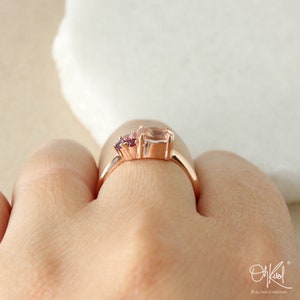 May include: A rose gold ring with a large oval-shaped pink gemstone and two smaller purple gemstones. The ring is on a person's finger.