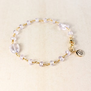 May include: A delicate gold chain bracelet with small, light purple beads and a gold charm with the letter "B" on it.