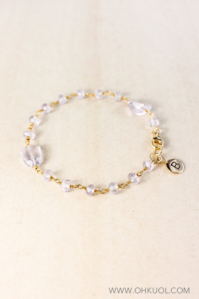 May include: A delicate gold chain bracelet with small, light purple beads and a gold charm with the letter "B" on it.