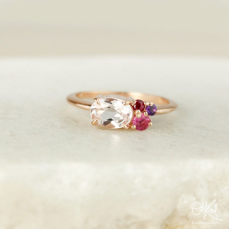 May include: A rose gold ring with a large oval-shaped clear gemstone and smaller pink, red, and purple gemstones.