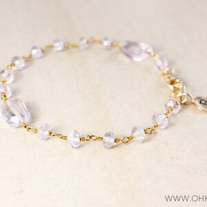 May include: A delicate gold chain bracelet with clear and light purple glass beads. The bracelet has a small charm with the letter "B" on it.