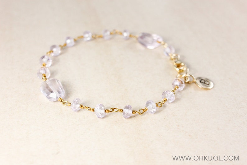 May include: A delicate gold chain bracelet with clear and light purple glass beads. The bracelet has a small charm with the letter "B" on it.