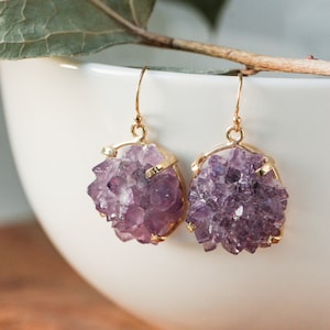 May include: A pair of amethyst earrings with gold-colored frames. The earrings feature clusters of purple crystals, suspended from gold-colored ear wires. The earrings are displayed against a blurred white background.