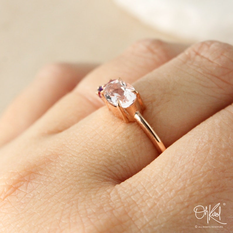 May include: A rose gold ring with a large oval-shaped clear gemstone and smaller pink and purple gemstones on the band.