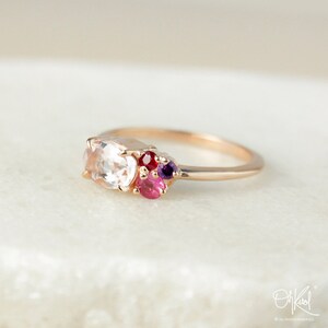 May include: A delicate rose gold ring with a cluster of colorful gemstones. The ring features a large oval-shaped white gemstone surrounded by smaller pink, red, and purple gemstones.