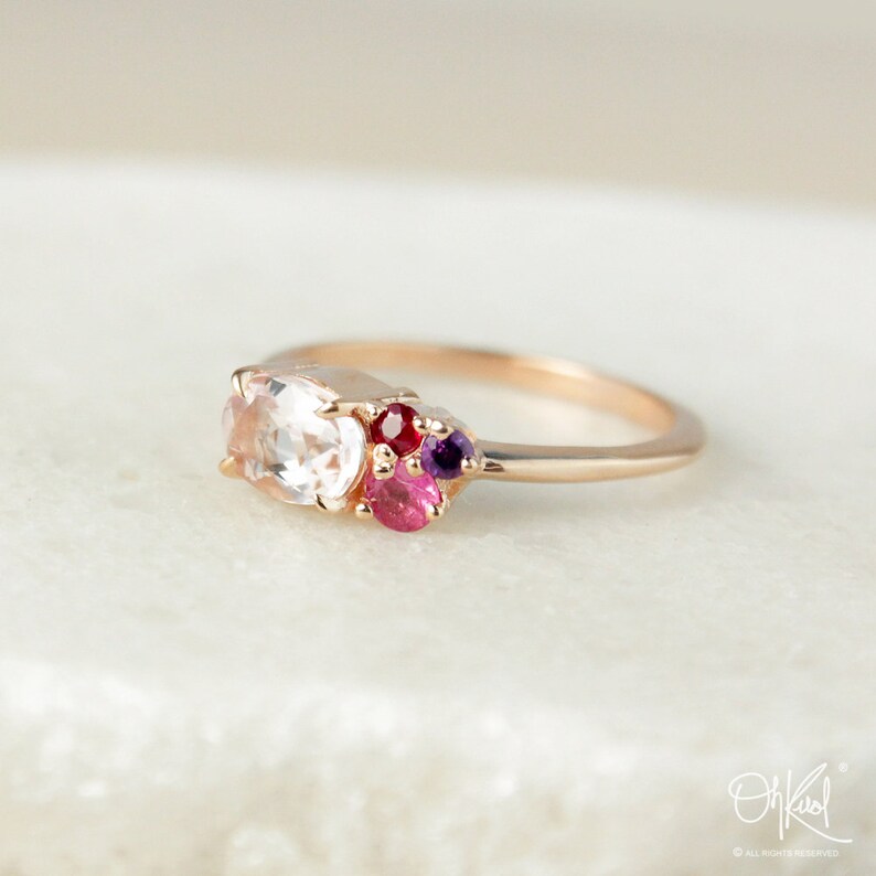 May include: A delicate rose gold ring with a cluster of colorful gemstones. The ring features a large oval-shaped white gemstone surrounded by smaller pink, red, and purple gemstones.