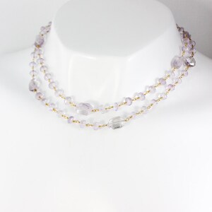May include: A delicate layered necklace with small, light purple beads and a larger, clear crystal bead. The necklace is made of gold chain and is displayed on a white mannequin.