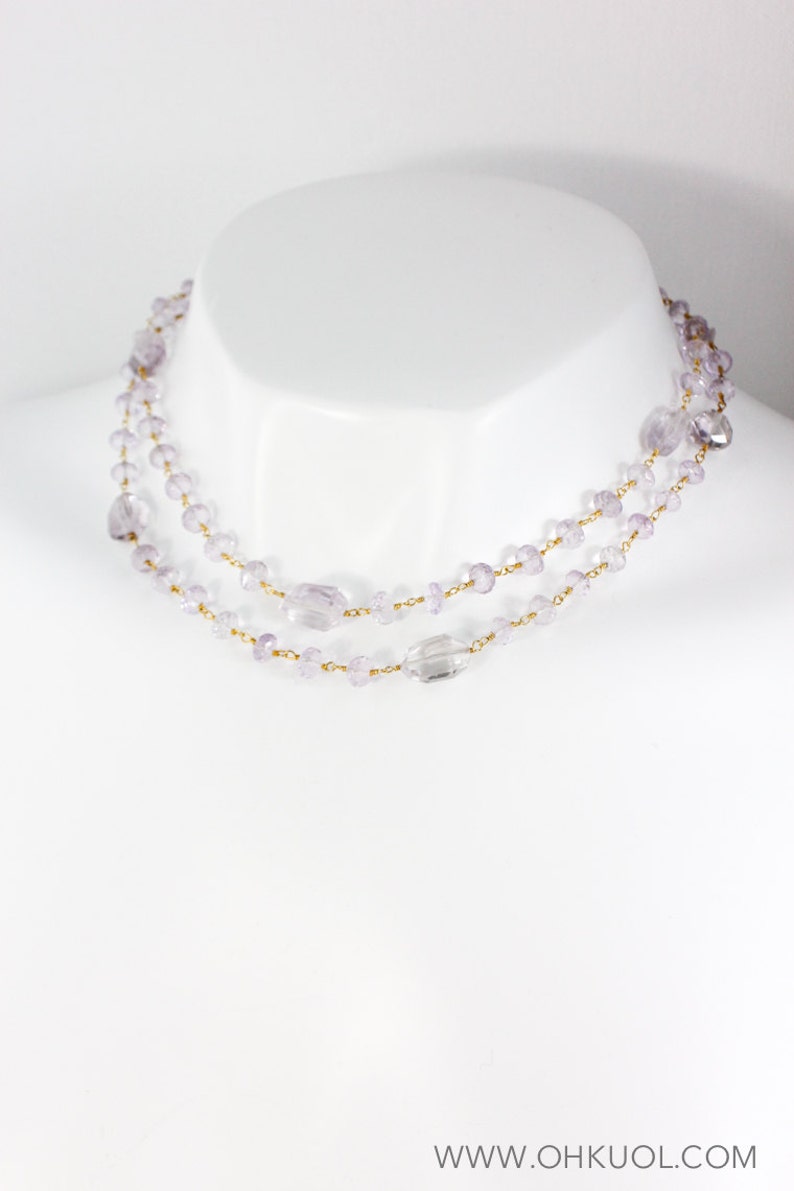 May include: A delicate layered necklace with small, light purple beads and a larger, clear crystal bead. The necklace is made of gold chain and is displayed on a white mannequin.