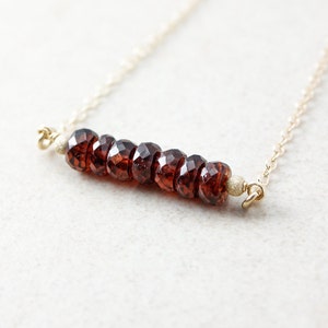 Pu&ograve; includere: Una collana a catena dorata con una barra di perle di granato sfaccettate. La collana &egrave; su uno sfondo bianco.