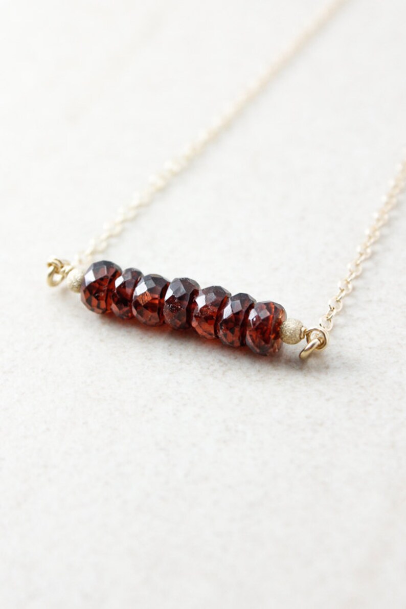 Pu&ograve; includere: Una collana a catena dorata con una barra di perle di granato sfaccettate. La collana &egrave; su uno sfondo bianco.