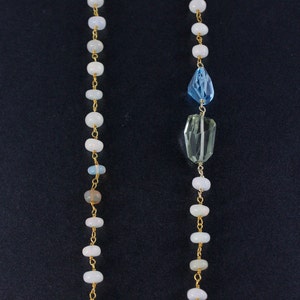 May include: A long gold chain necklace with white beads and two gemstone pendants. The top pendant is a light blue teardrop shape and the bottom pendant is a light green rectangular shape.