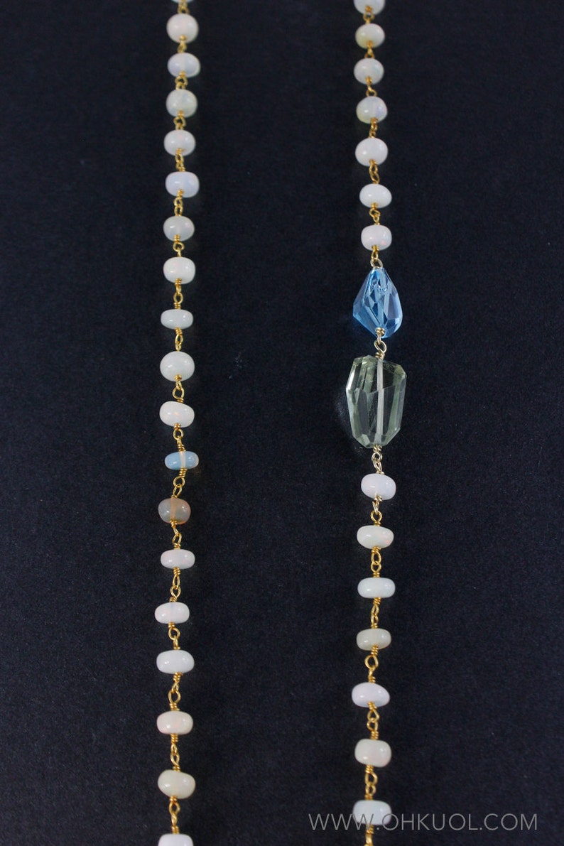 May include: A long gold chain necklace with white beads and two gemstone pendants. The top pendant is a light blue teardrop shape and the bottom pendant is a light green rectangular shape.