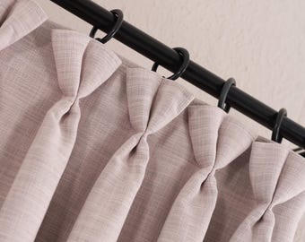 May include: A light mauve curtain with a textured fabric finish featuring pinch pleats at the top. The curtain is suspended from a black metal rod by circular black rings, creating uniform vertical folds that drape downward from the rod.