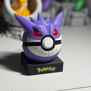 May include: A purple Gengar figure with red eyes and a white and black Poké Ball design. The figure sits on a black base with the word "Pokémon" in gold lettering. Other Pokémon figures are visible in the background.