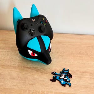 May include: A black Xbox controller rests in a blue and black Lucario-themed controller holder. A pixelated Lucario figure is next to the holder on a wooden surface. The holder has blue ears and red eyes.