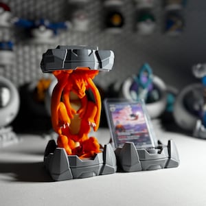 May include: A 3D-printed orange Charizard figure is held within a gray, rocky-textured display stand. A trading card is displayed in a matching stand. Other figures are visible in the background.