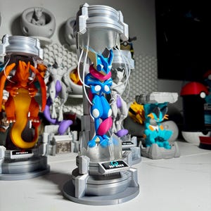 May include: A blue and pink Greninja figurine suspended inside a silver cylindrical containment chamber with a clear plastic tube. The base features a small black plaque with the text No. 0658 and a blue water droplet icon printed on the front.
