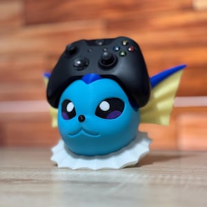 May include: A blue and yellow Pokemon character with a white base holding a black Xbox controller.