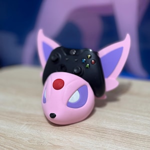 May include: A pink and purple Pokemon-themed controller stand with a black Xbox controller in the stand.