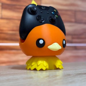 May include: A black and orange video game controller stand shaped like a cartoon bird. The stand has a yellow beak and yellow feet.
