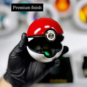 May include: A spherical red and white ball-shaped container with a black band and a central silver circular button. The object features a smooth, glossy surface and includes the text Premium finish. The interior is dark with a green light.