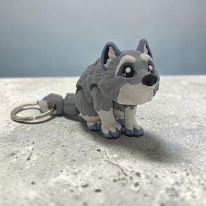 May include: A gray and white wolf figurine keychain with a stylized, cartoon-like appearance. The wolf has large, dark eyes, a black nose, and textured fur detailing. A silver-toned metal split ring is attached to the back of the wolf figure.