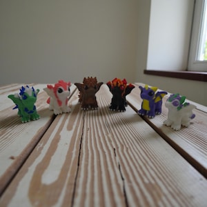 May include: A selection of colourful dragon figurines. The figurines are in various colours, including green, red, brown, black, purple, and white. Each dragon has unique features, such as wings, horns, and spikes. They are arranged on a light-coloured wooden surface.