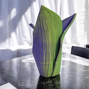 May include: A green and purple ceramic vase with a unique, folded design. The vase is made of a textured material and has a modern, abstract look.