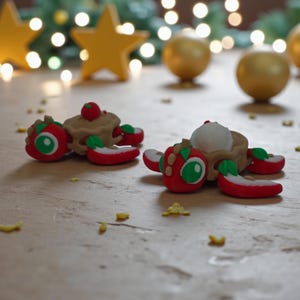 May include: Two whimsical, miniature food sculptures. One features a tan pie topped with a red cherry, surrounded by red apple slices. The other has a white scoop of ice cream in a tan bowl, with red apple slices. Green and gold Christmas decorations are in the background.